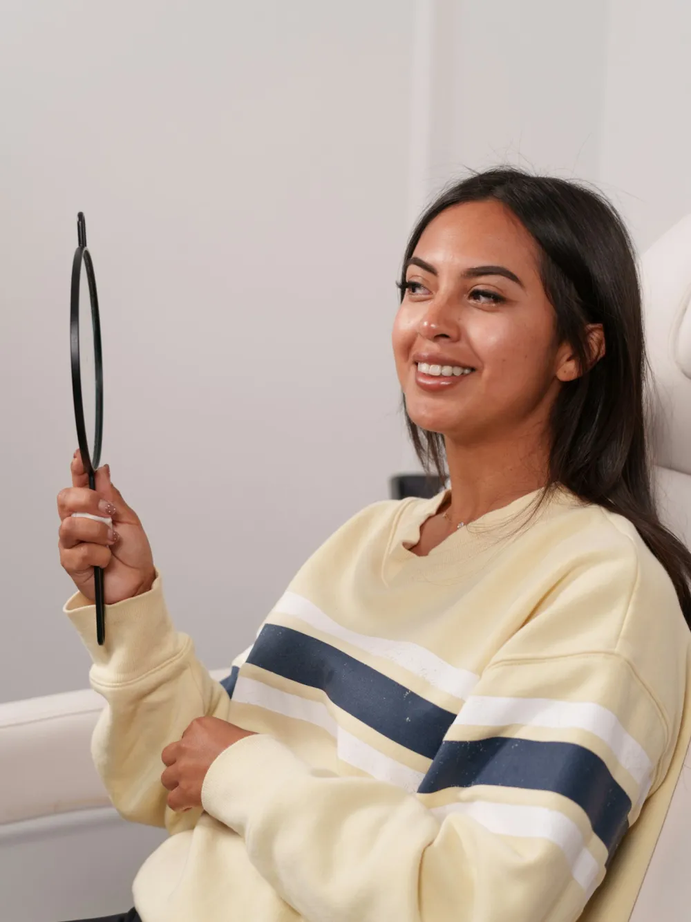 Woman looking at her lips in a hand mirror while discussing goals during a cosmetic consultation